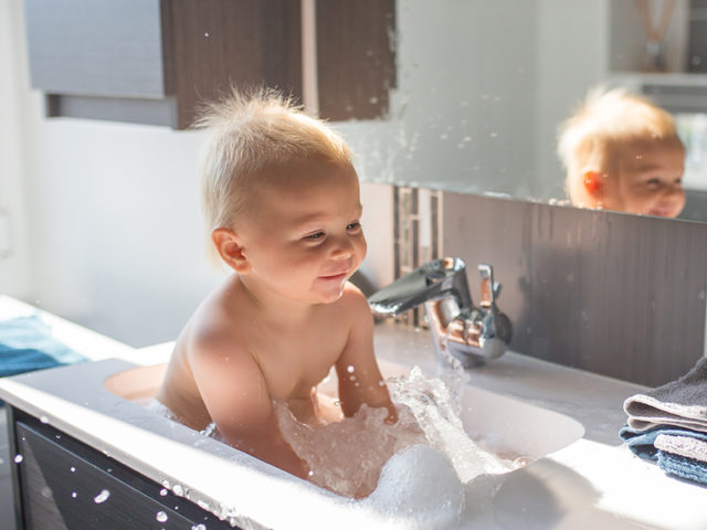 Baby taking bath in sink. Child playing with foam and soap bubbles in sunny bathroom with window. Little boy bathing. Water fun for kids. Hygiene and skin care for children. Bath room interior
