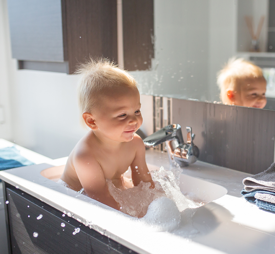 Baby taking bath in sink. Child playing with foam and soap bubbles in sunny bathroom with window. Little boy bathing. Water fun for kids. Hygiene and skin care for children. Bath room interior